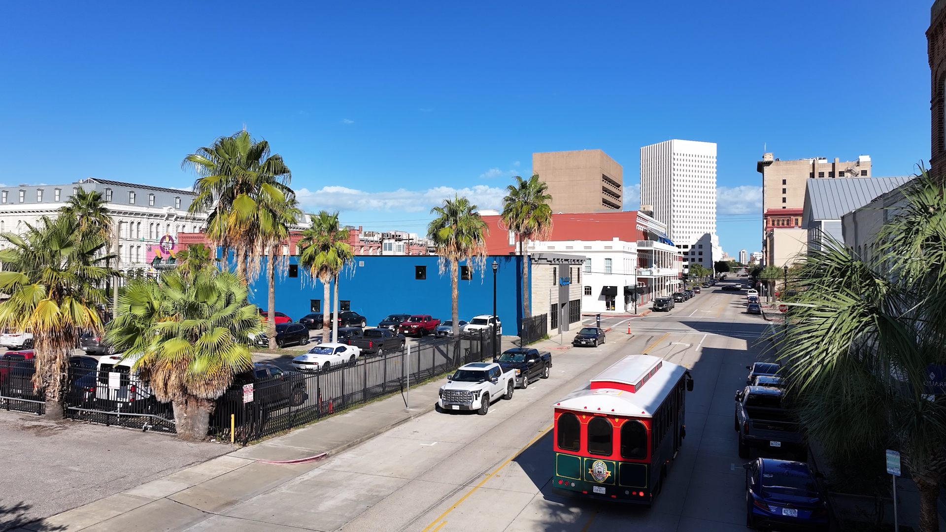 A Galveston trolley moving through downtown among palms, parked cars, sidewalks and buildings